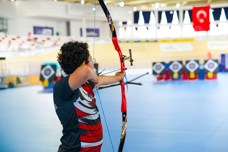 Sporcu kırmızı olimpik yayla ankrajda; arkada Türk bayrağı ve hedef sırası