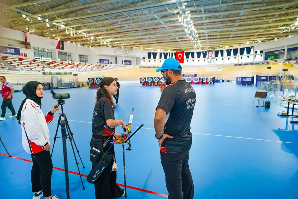 Antrenör ve sporcular atış hattının gerisinde; tripod üzerinde gözlem dürbünü