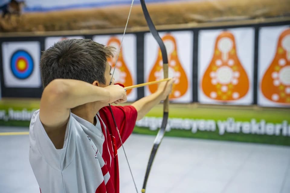 Genç sporcu salonda geleneksel yayla nişan alıyor; arkada puta hedefleri