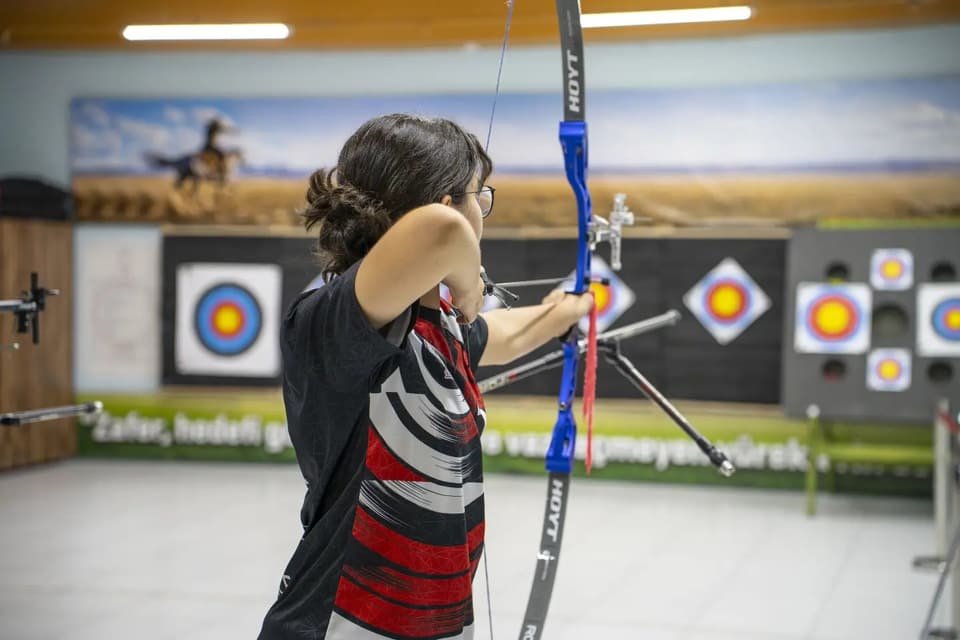 Sporcu salonda mavi olimpik yayla çekiş yapıyor; duvarda renkli hedefler